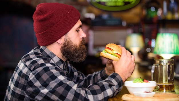 Hipster rilassante al pub. Il pub è un luogo rilassante dove bere e rilassarsi. L'uomo con la barba beve birra mangia il menu dell'hamburger. Goditi il pasto al pub. L'uomo barbuto brutale hipster si siede al bancone del bar. Spuntino ipercalorico.