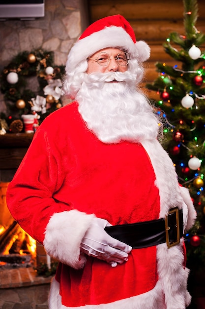Ho ho ho! Babbo Natale allegro che si aggiusta la cintura e sorride con il camino e l'albero di Natale sullo sfondo