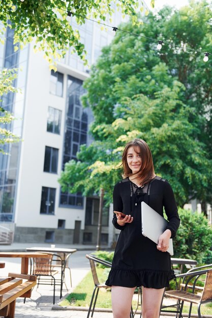 Holding laptop e telefono Ragazza felice in gonna nera all'aperto in città vicino a alberi verdi e contro la costruzione di affari