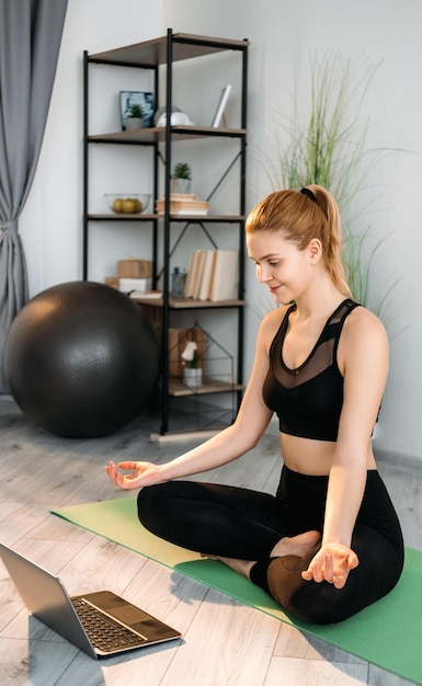 Home yoga Benessere online Mente corpo equilibrio Donna rilassata che guarda la sessione di terapia di allenamento sul laptop seduto a gambe incrociate sul pavimento facendo esercizi calmanti in interni moderni