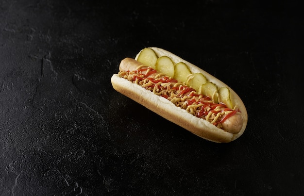 Hot dog con cetrioli sottaceto, cipolle fritte, ketchup e senape su sfondo nero.