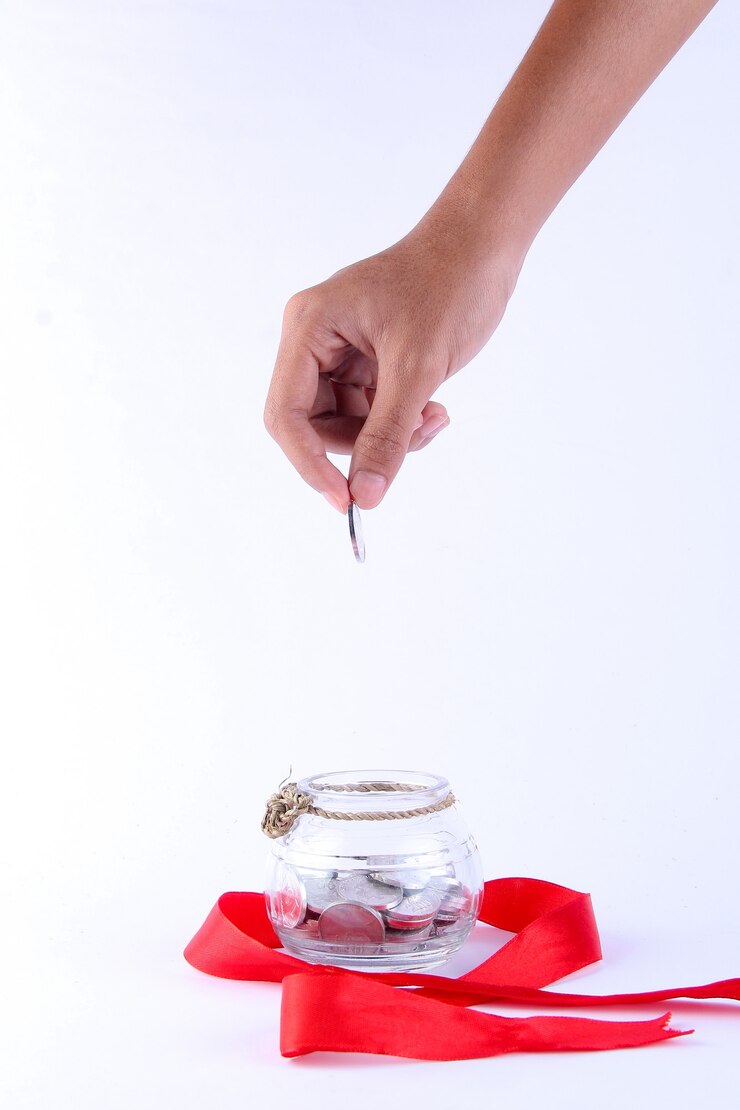 Photo human hand holding a coin to put in a transparent jar decorated with a red ribbon around it