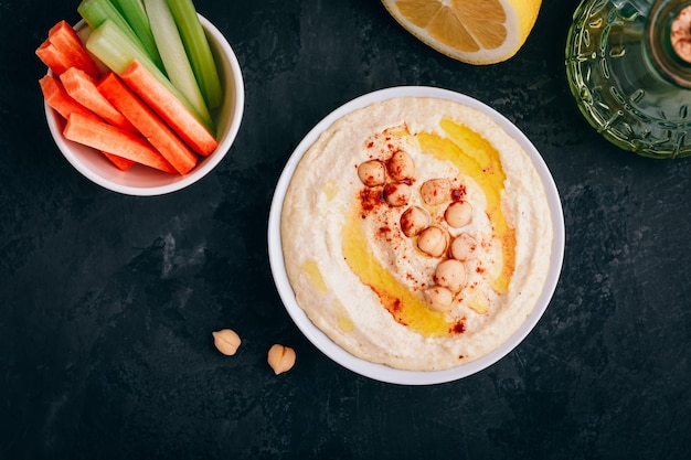 Hummus con olio d'oliva e ceci in una ciotola con bastoncini vegetali su sfondo di pietra scura Vista dall'alto con spazio per la copia