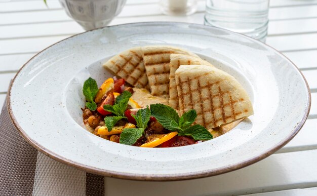 Hummus con pomodori ed erbe aromatiche in piatto bianco con pita sul tavolo e bicchiere d'acqua cafe cibo vegano