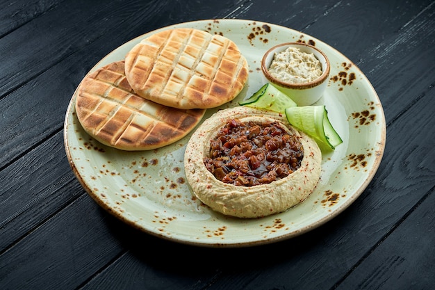 Hummus di ceci con verdure al forno e olio d'oliva servito con pita al forno in un piatto su uno sfondo di legno scuro.