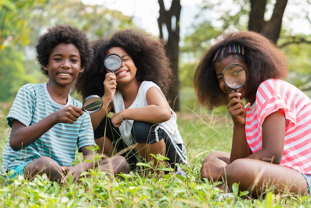 I bambini afroamericani che si siedono nell'erba e che guardano attraverso la lente d'ingrandimento tra imparano oltre l'aula. Concetto di educazione all'aperto.