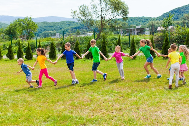 I bambini corrono tenendosi per mano all'aperto. Ragazze e ragazzi si divertono insieme sul prato verde, giorno d'estate