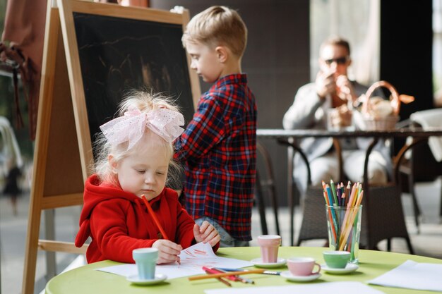 I bambini disegnano. La ragazza bionda si siede a un tavolo, un ragazzo si trova vicino a una lavagna.