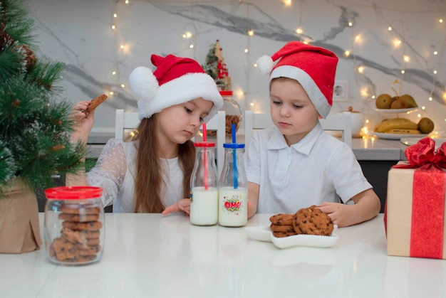 I bambini felici con i cappelli di Babbo Natale si raccontano storie divertenti. Sorella e fratello bevono latte, mangiano biscotti vicino a casa in cucina con decorazioni natalizie alla vigilia di Natale a casa. Messa a fuoco selettiva.