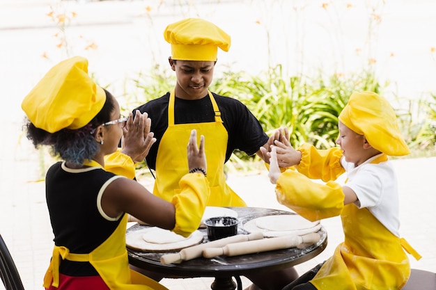 I bambini multirazziali cuochi giocano con la farina per l'impasto e si divertono Bambini multinazionali con cappello da cuoco e grembiule giallo che cucinano uniforme all'aperto per la panetteria
