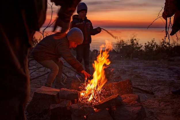 I bambini si siedono vicino al fuoco sulla spiaggia autunnale dopo il tramonto si scaldano le mani si divertono insieme all'esterno