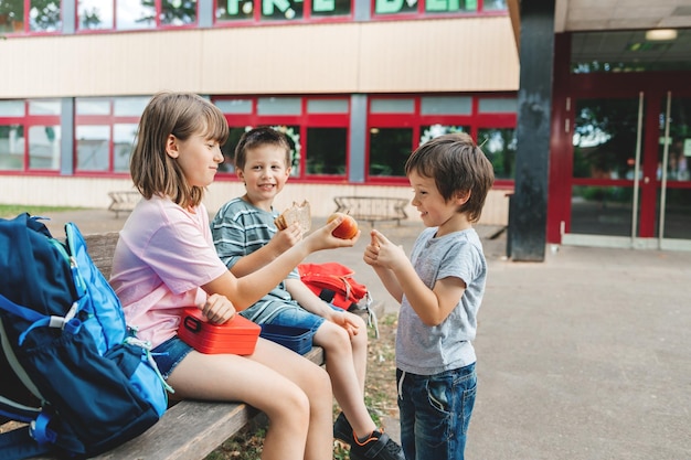 I bambini siedono su una panchina nel cortile della scuola e mangiano mele e panini durante la pausa