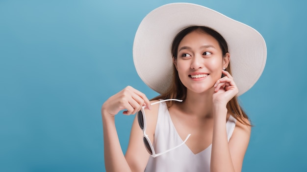 I bei blogger sorridenti asiatici indossano un ampio cappello e giudicano gli occhiali da sole isolati nel fondo blu