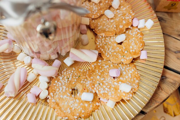 I biscotti con granelli di zucchero giacciono su un piatto d'oro, marshmello, tavolo di legno, il concetto di Natale sono sparsi in giro.