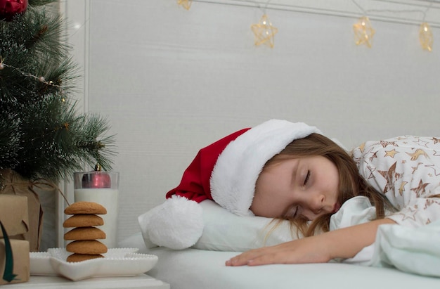 I biscotti di Natale e un bicchiere di latte sono una delizia tradizionale. Un'affascinante bambina si è addormentata sul letto vicino al comodino con un regalo per Babbo Natale. Il concetto del nuovo anno. Parenti