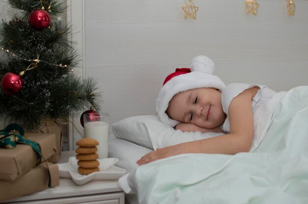 I biscotti di Natale e un bicchiere di latte sono una delizia tradizionale. Un bambino affascinante si è addormentato sul letto vicino al comodino con un regalo per Babbo Natale. Il concetto del nuovo anno. Parenti.