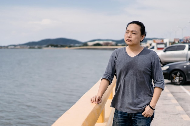 I capelli lunghi maschili asiatici con una maglietta grigio scuro si ergono sulla sposa accanto alla strada e non vedono l'ora che arrivi il mare nel periodo pomeridiano