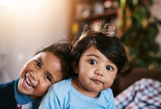 I fratelli si uniscono Ritratto di un bambino allegro e di sua sorella maggiore che si rilassano a casa mentre guardano nella fotocamera all'interno durante il giorno