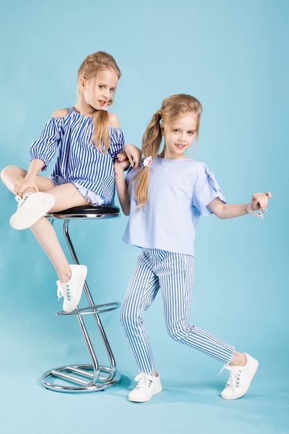 I gemelli delle ragazze in vestiti blu-chiaro stanno posando vicino ad uno sgabello da bar su un blu.