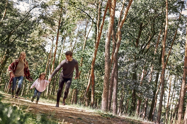 I genitori felici con la loro figlia si tengono per mano e corrono sul sentiero nel bosco durante la passeggiata nella foresta autunnale. Stagione fredda, famiglia bella e felice