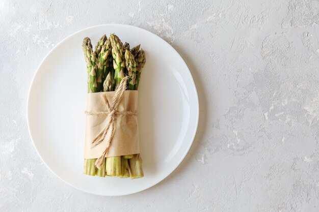 I germogli di asparagi sono legati con una corda di iuta in un piatto bianco. L'asparago fresco è avvolto in carta su uno sfondo di cemento con una copia dello spazio.