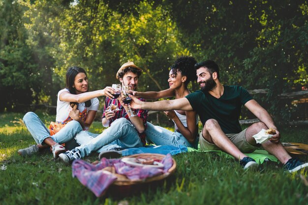 I giovani brindano al picnic Gruppo di amici che tintinnano bicchieri di vino e mangiano panini seduti sull'erba