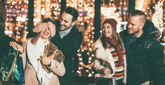 I giovani felici quattro amici nello shopping passano davanti al centro commerciale di vetrine e l'uomo tiene gli occhi della sua ragazza coperti mentre lei fa un regalo.