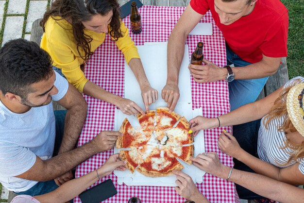 I giovani felici si precipitano sulla loro fetta di pizza appena consegnata tavolo da picnic con tovaglia cartone pizza e bottiglie di birra tipico concetto di festa alimentare italiana e incontri felici spensierati