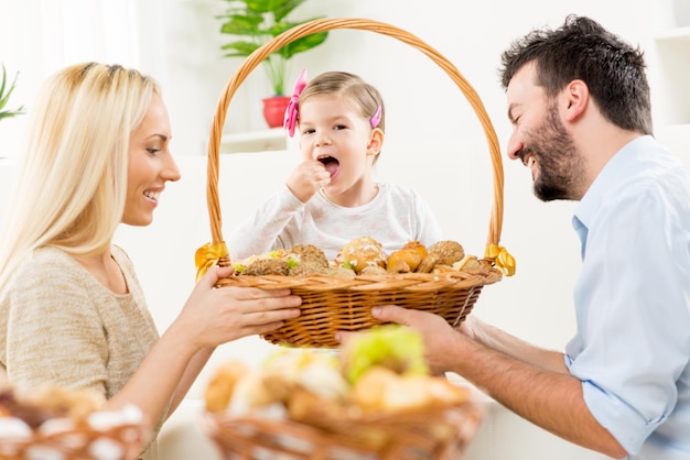 I giovani genitori si inginocchiano davanti alla loro piccola figlia con un sorriso guardando i pasticcini da un grande cesto intrecciato che insieme tengono nelle loro mani.