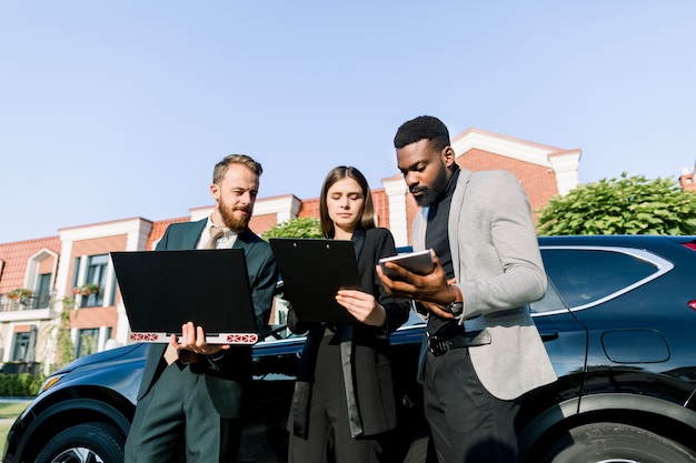 I giovani soci d'affari di fiducia, l'uomo africano e l'uomo e la donna caucasici, stanno lavorando insieme utilizzando tablet e laptop prima del viaggio d'affari in piedi vicino all'automobile nera all'aperto.