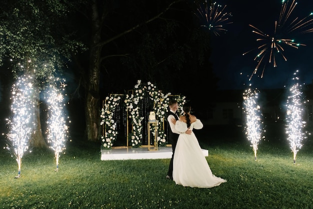 I giovani sposi stanno vicino all'arco delle nozze di notte con le luci del fumo e le sagome dei fuochi d'artificio degli sposi novelli