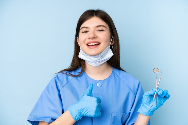 I giovani strumenti di tenuta della donna del dentista sopra la parete blu isolata che dà pollici aumentano il gesto