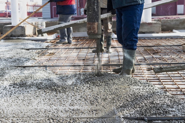 I lavoratori dei costruttori versano il pavimento in cemento nell'officina industriale Gambe negli stivali in cemento Presentazione del cemento per il getto del pavimento Opere monolitiche in cemento