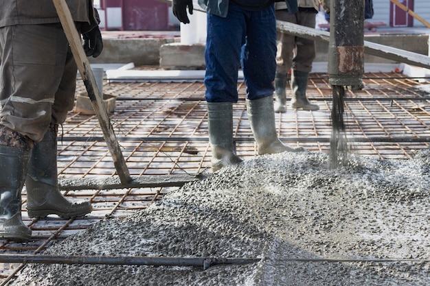 I lavoratori dei costruttori versano il pavimento in cemento nell'officina industriale Gambe negli stivali in cemento Presentazione del cemento per il getto del pavimento Opere monolitiche in cemento
