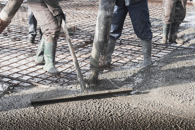 I lavoratori dei costruttori versano il pavimento in cemento nell'officina industriale Gambe negli stivali in cemento Presentazione del cemento per la colata del pavimento Opere monolitiche in cemento