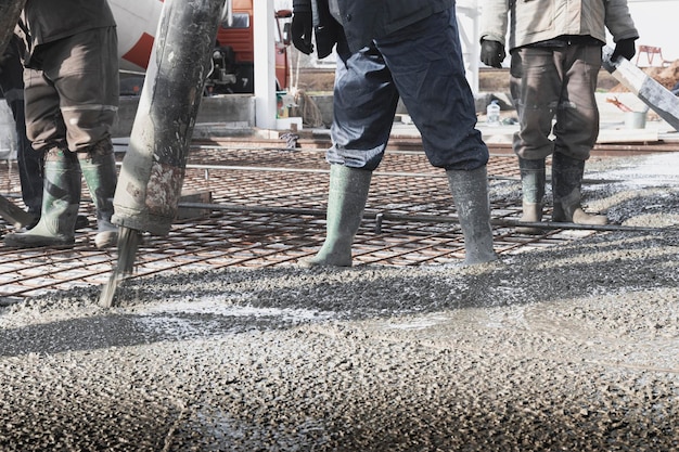 I lavoratori dei costruttori versano il pavimento in cemento nell'officina industriale Gambe negli stivali in cemento Presentazione del cemento per la colata del pavimento Opere monolitiche in cemento