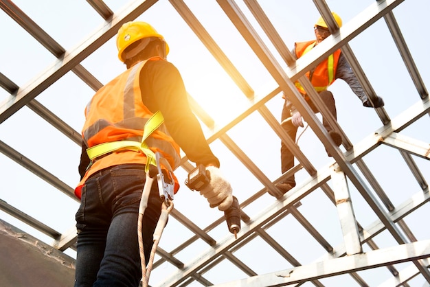 I lavoratori edili indossano cinghie di sicurezza mentre lavorano sulla struttura del tetto dell'edificio in un cantiere edile Roofer utilizzando una pistola sparachiodi pneumatica