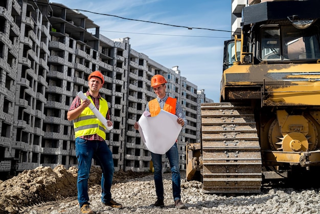 I lavoratori sono forti in tuta e caschi con un piano in mano in piedi vicino al nuovo concetto di costruzione dell'edificio