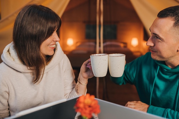 I liberi professionisti felici della coppia della famiglia bevono il caffè mentre lavorano il computer portatile sull'accogliente tenda glamping nella sera d'estate Tenda da campeggio di lusso per le vacanze all'aperto e il concetto di stile di vita di vacanza