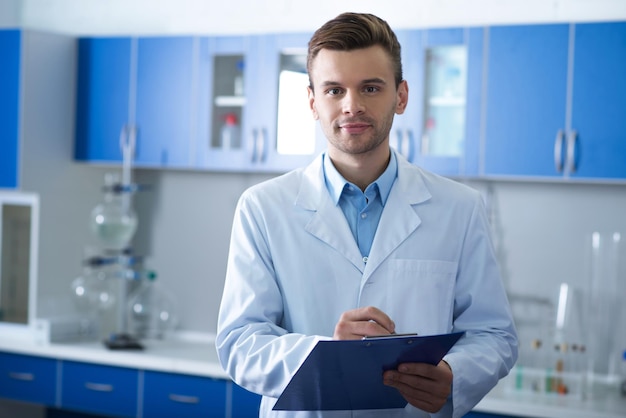 I migliori datori di lavoro Scienziato adulto bello e sicuro di sé in piedi in laboratorio guardando dritto e tenendo il suo taccuino