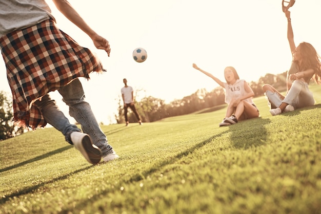 I migliori fan! Due giovani donne sedute sull'erba e gesticolano mentre i loro amici maschi giocano a calcio all'aperto