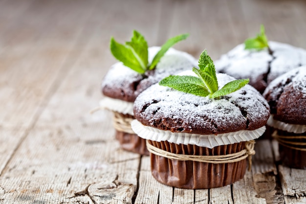 I muffin scuri del cioccolato fresco con lo zucchero in polvere e la menta coprono di foglie sul fondo di legno rustico della tavola.