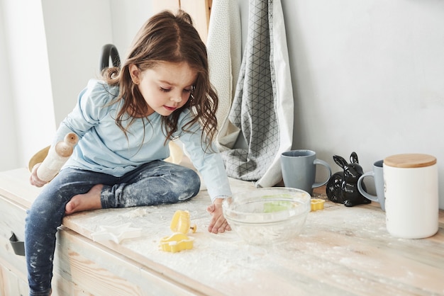 I pantaloni sono sporchi. Foto di una bambina graziosa che si siede sul tavolo della cucina e gioca con la farina.