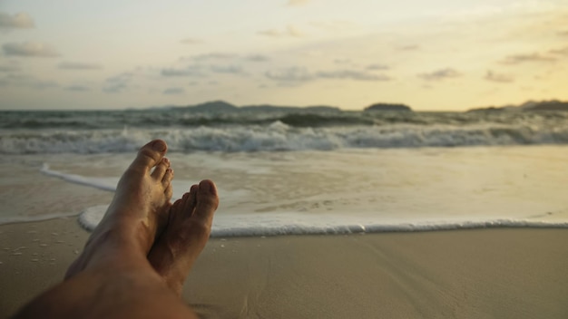 I piedi dell'uomo rilassati sono sdraiati sulla spiaggia sabbiosa e bagnati dall'acqua e dalla schiuma dell'oceano