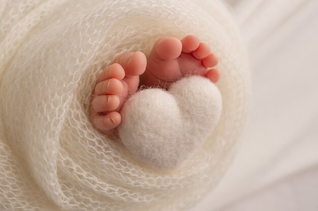 I piedi di un neonato sono avvolti in una coperta a maglia. Le dita di un neonato tengono in mano un cuore a maglia bianca. Foto di alta qualità