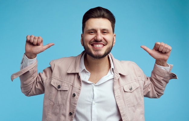 I punti maschii caucasici con entrambi i pollici a se stesso hanno un ampio sorriso in piedi su sfondo blu