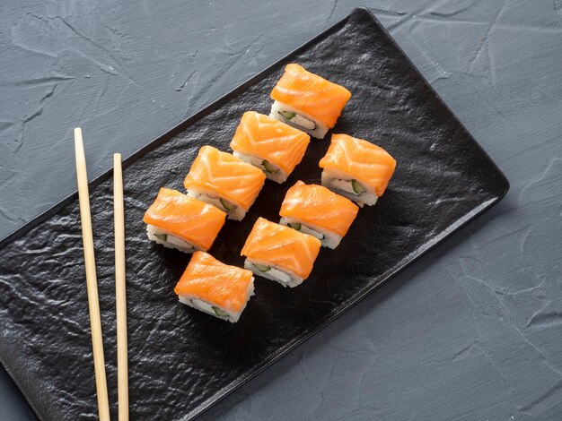 I rotoli di sushi di Filadelfia su un supporto nero strutturato del piatto su un fondo grigio. Ci sono bacchette di bambù nelle vicinanze. Vista dall'alto, piatto. cucina giapponese