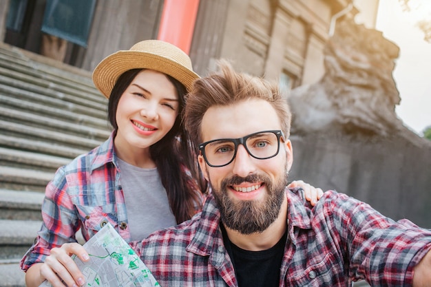 I turisti simpatici e positivi sorridono e posano sulla macchina fotografica. Il giovane lo tiene. Inoltre ha una mappa in mano. La giovane donna sta dietro di lui e sorride anche. Sono molto positivi.