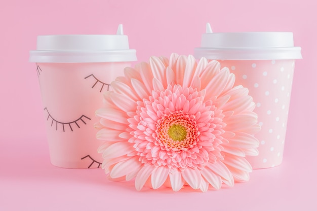 I vetri di caffè e la gerbera da asporto fioriscono su fondo pastello rosa.