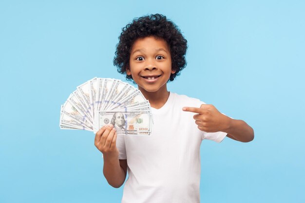 I39m ricco Gioioso fortunato ragazzino ricco con i capelli ricci che puntano al fan di banconote in dollari che si vantano soldi e guardando la fotocamera con eccitato sorriso a trentadue denti in studio indoor girato sfondo blu